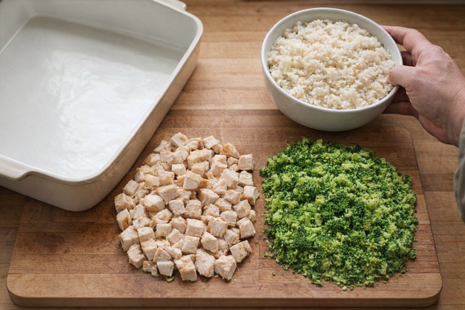 An overhead shot of a cutting board with diced chicken, chopped broccoli, and a bowl of cooked rice, next to a 9x13 baking dish